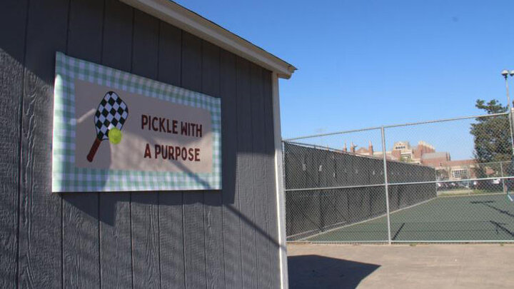 Sign reading "Pickle With a Purpose" on a shed, next to a fenced pickleball court under clear skies.