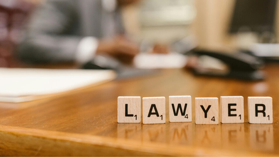 Scrabble tiles spelling "LAWYER" on a wooden desk, blurred figure in background.