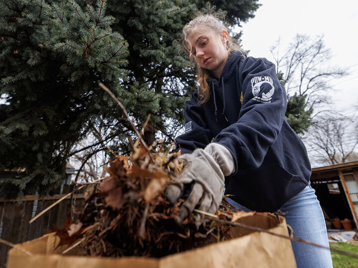 Person wearing gloves placing dried leaves into a brown bag outdoors near trees.