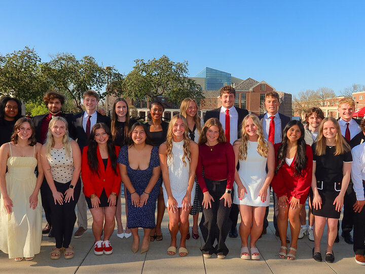 FCLA Class of 2025. Group of young adults in formal attire posing outdoors on a sunny day, surrounded by trees and buildings.